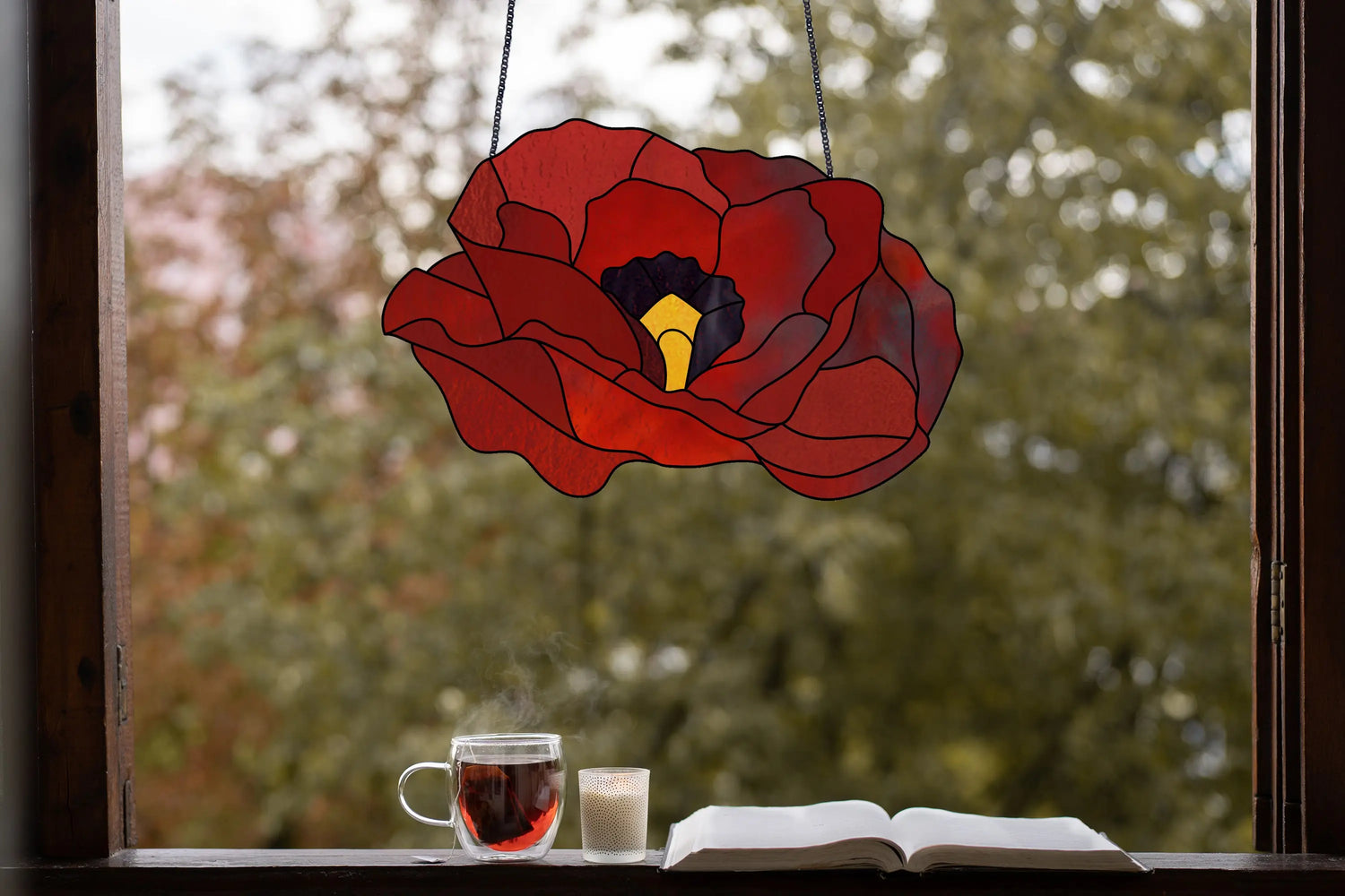 A red poppy-shaped stained glass art hangs in a window. Below, theres a steaming cup of tea, a lit candle, and an open book. Outside, trees with blurred green foliage are visible.