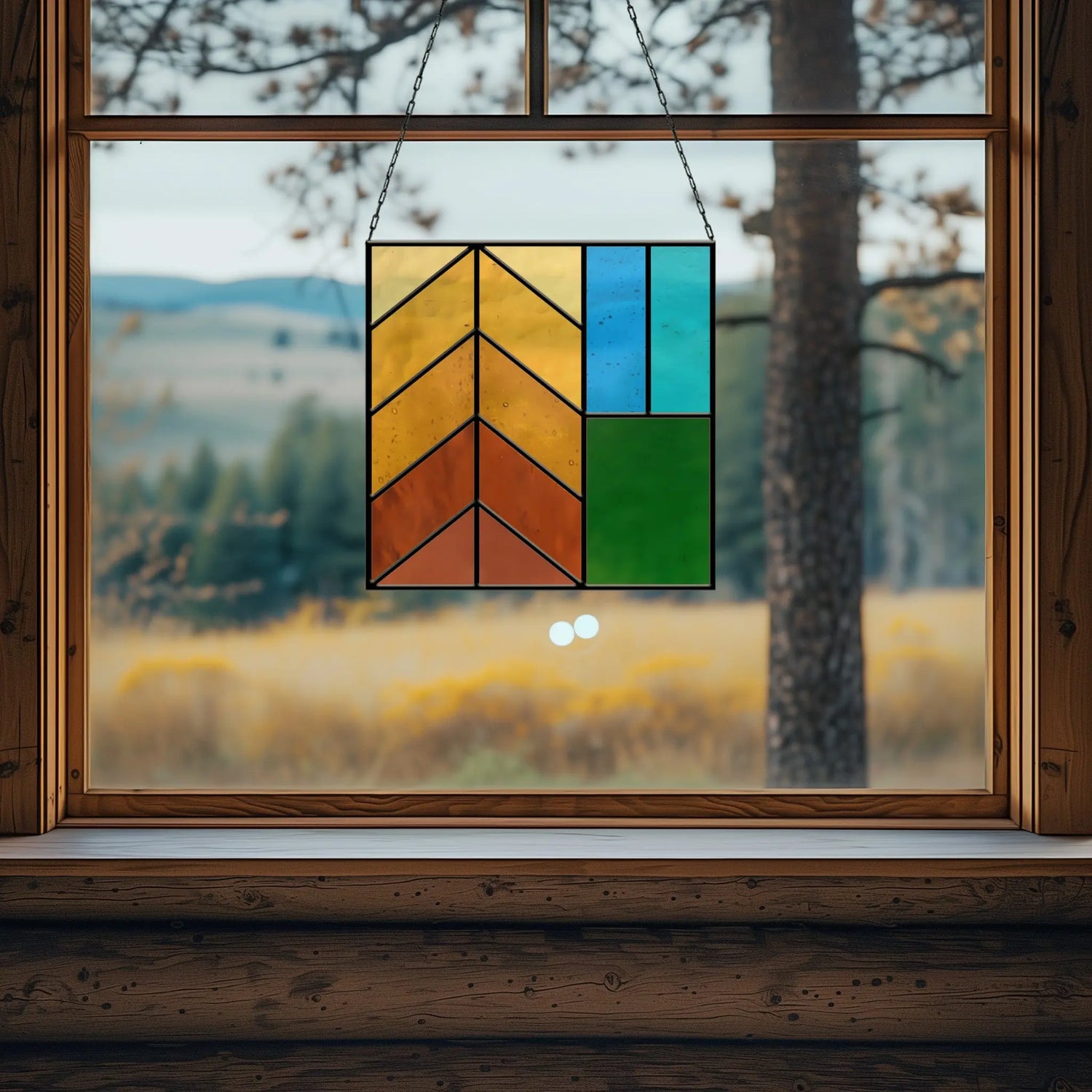 Stained glass art with geometric patterns in warm colors hangs in a rustic window. The background features a blurred landscape of trees and hills under a cloudy sky.