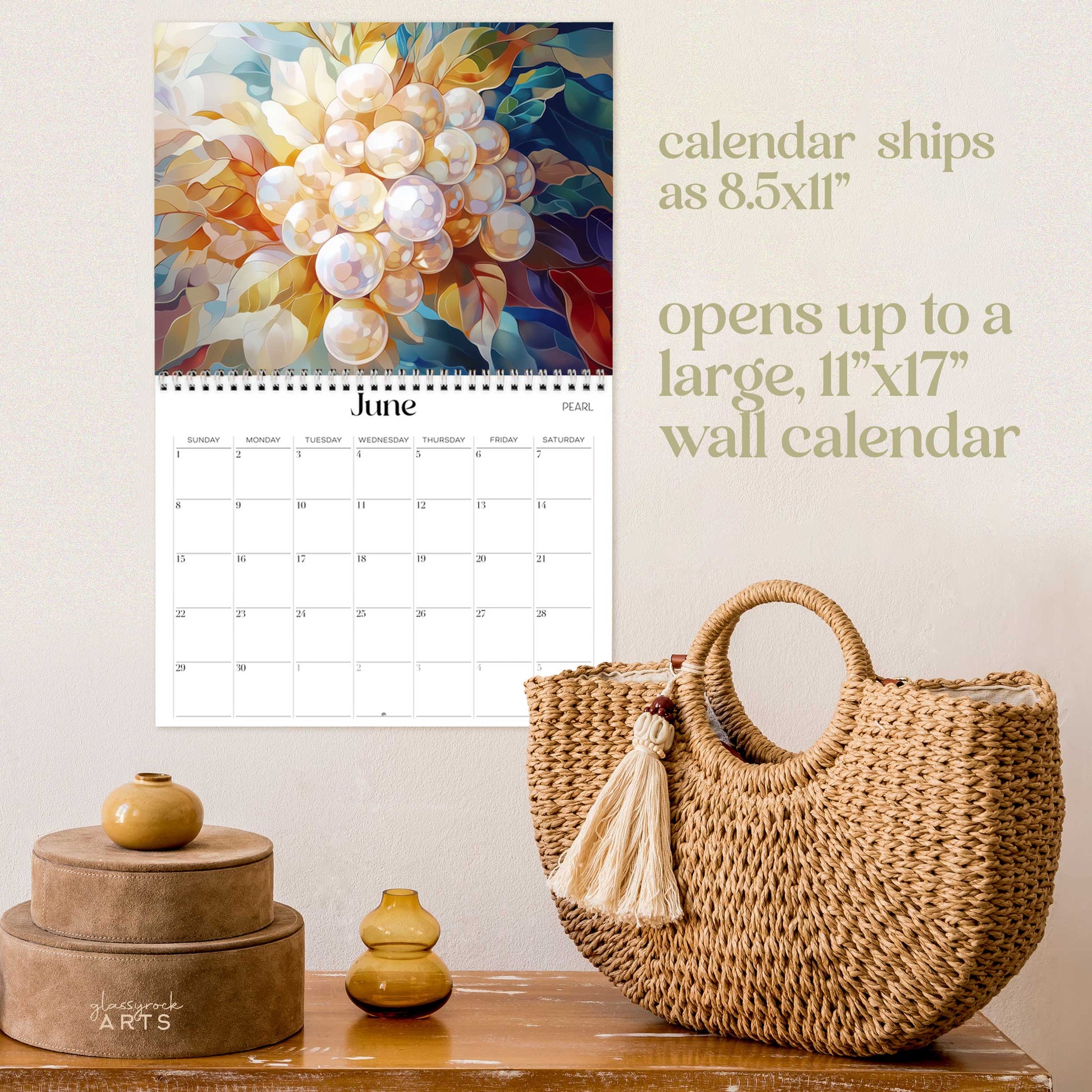 The birthstones calendar, a 2025 monthly wall calendar with gem-themed original artwork, on a wall above a wood table with a wicker purse. The calendar is opened to June showing pearls.