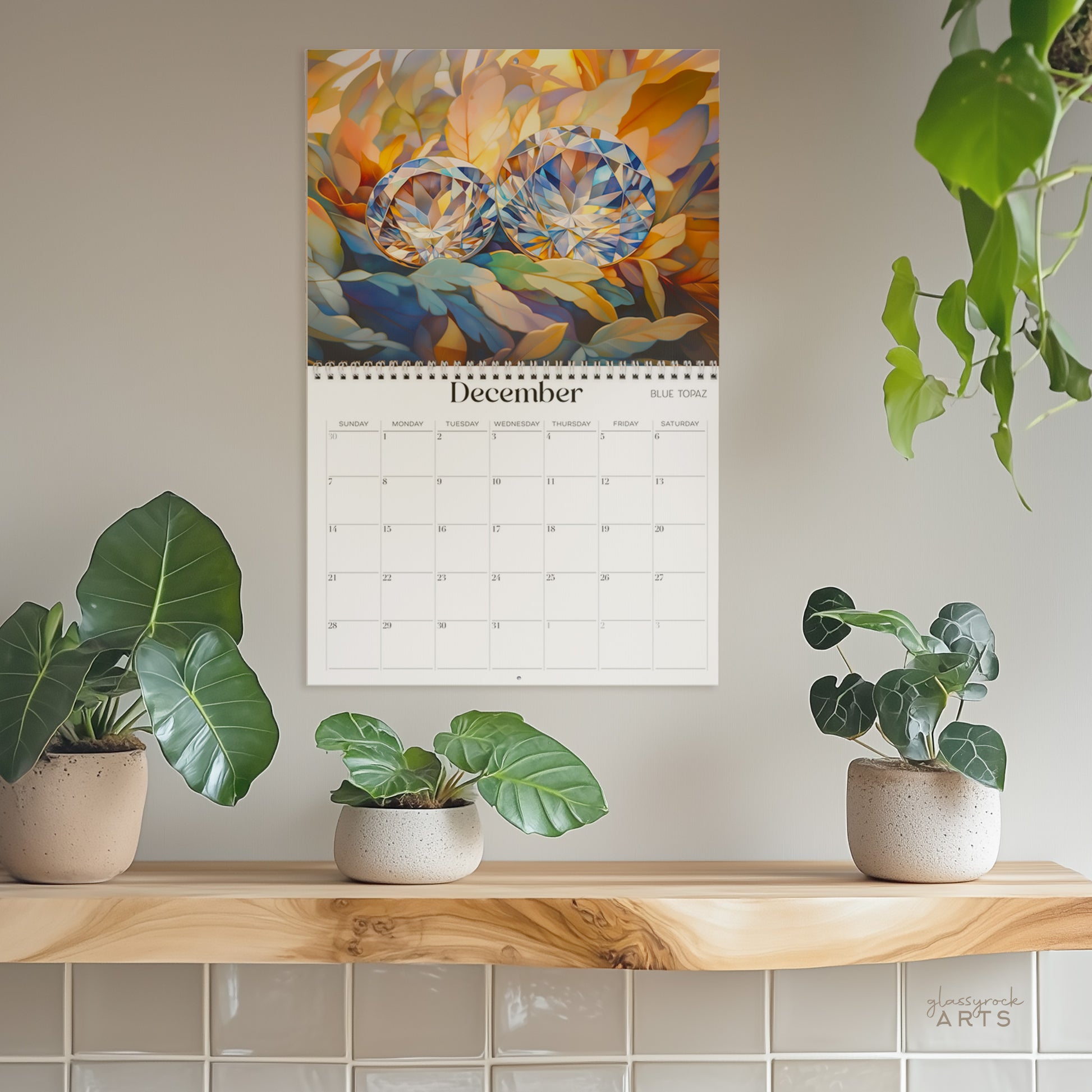 The birthstones calendar, a 2025 monthly wall calendar with gem-themed original artwork, hanging on a wall surrounded by plants. The December page shows blue topaz.