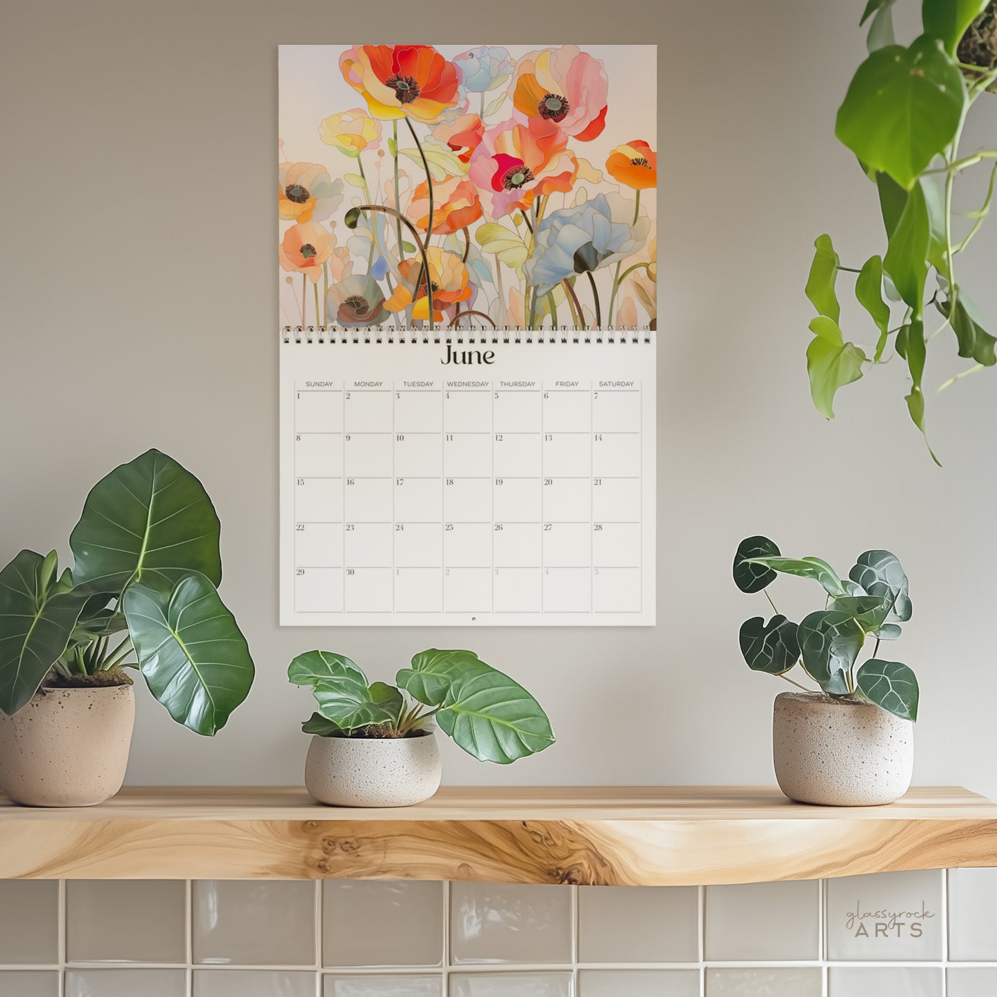 The poppies calendar, a 2025 monthly wall calendar with poppy-themed original artwork, hanging on a wall surrounded by plants. The June pages shows colorful poppies.