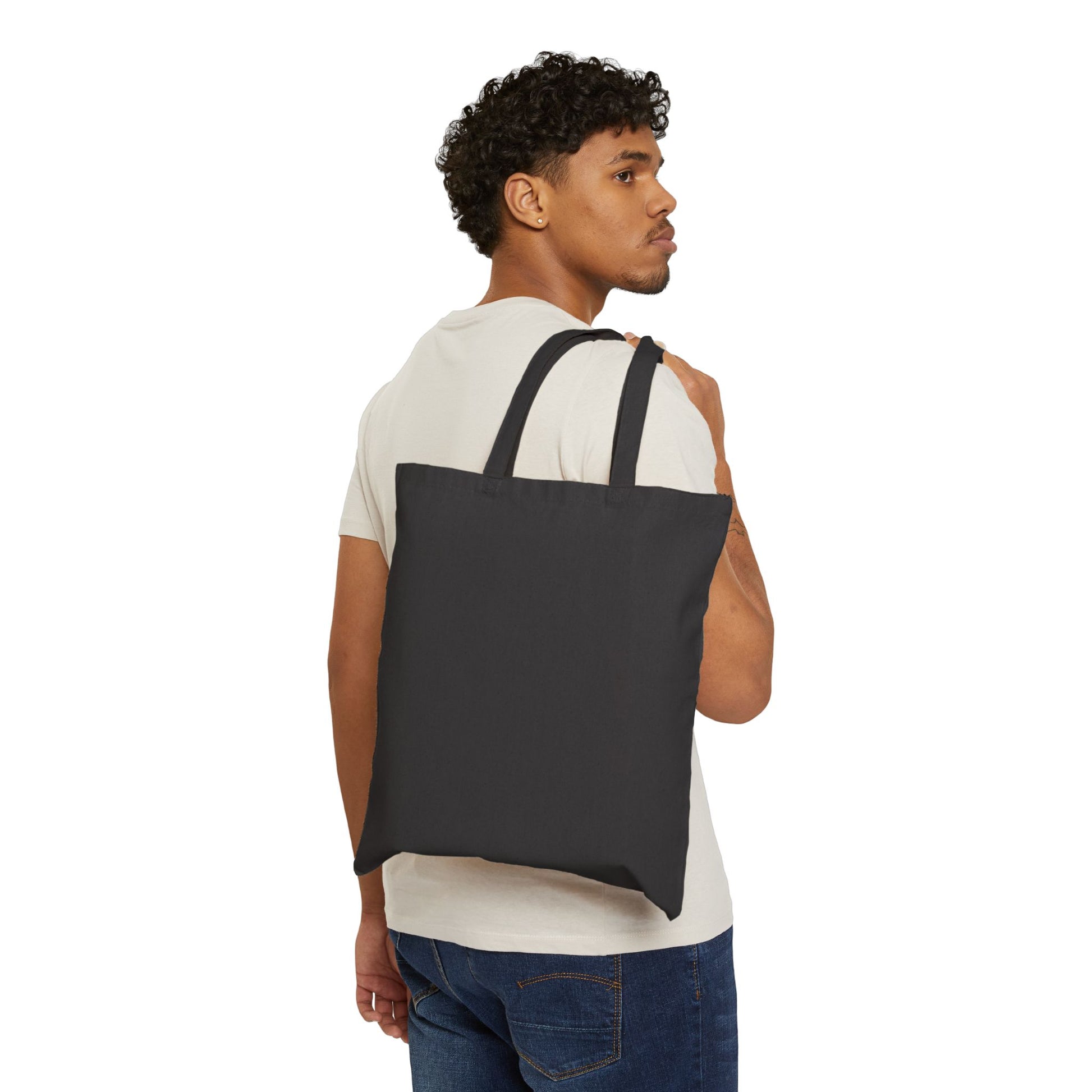 A man with curly hair, dressed in a white t-shirt and jeans, carries the Stained Glass Cactus Teardrop Cotton Canvas Tote Bag over his shoulder. The black tote showcases an original artwork inspired by stained glass design. He is looking to the side against a white background.