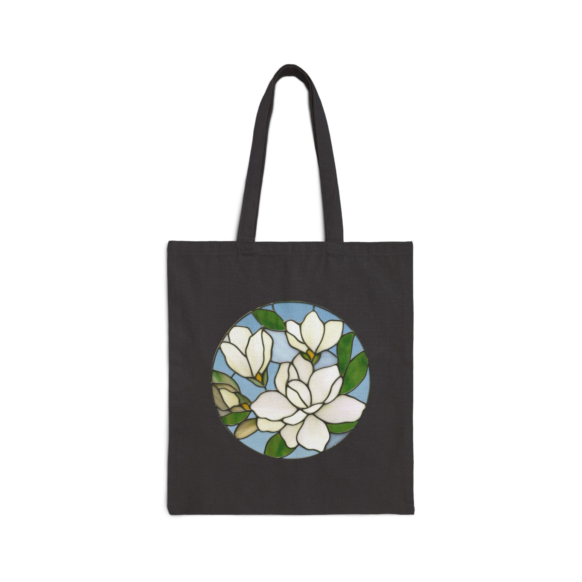 The Stained Glass Magnolia Cotton Canvas Tote Bag is a 100% cotton canvas tote bag showcasing original artwork of white magnolia flowers and green leaves, arranged in a circular, stained glass design on the front.