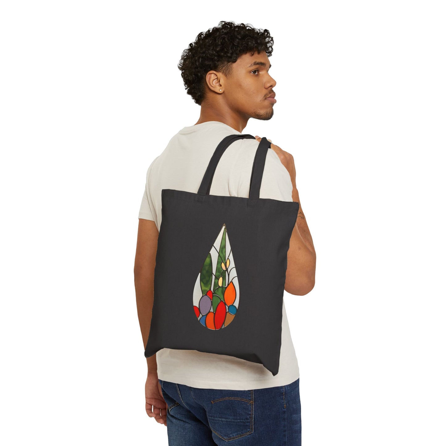 A man in a white t-shirt and jeans carries over his shoulder a black Stained Glass Cactus Teardrop Cotton Canvas Tote Bag featuring a colorful design. He stands against a plain white background, looking over his shoulder.