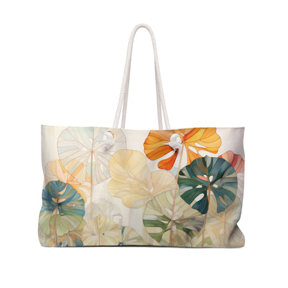 The Stained Glass Monstera Oversized Tote Bag displays a striking artistic print of overlapping tropical leaves in orange, blue, green, and beige shades with two rope handles, creating a colorful nature-inspired pattern.