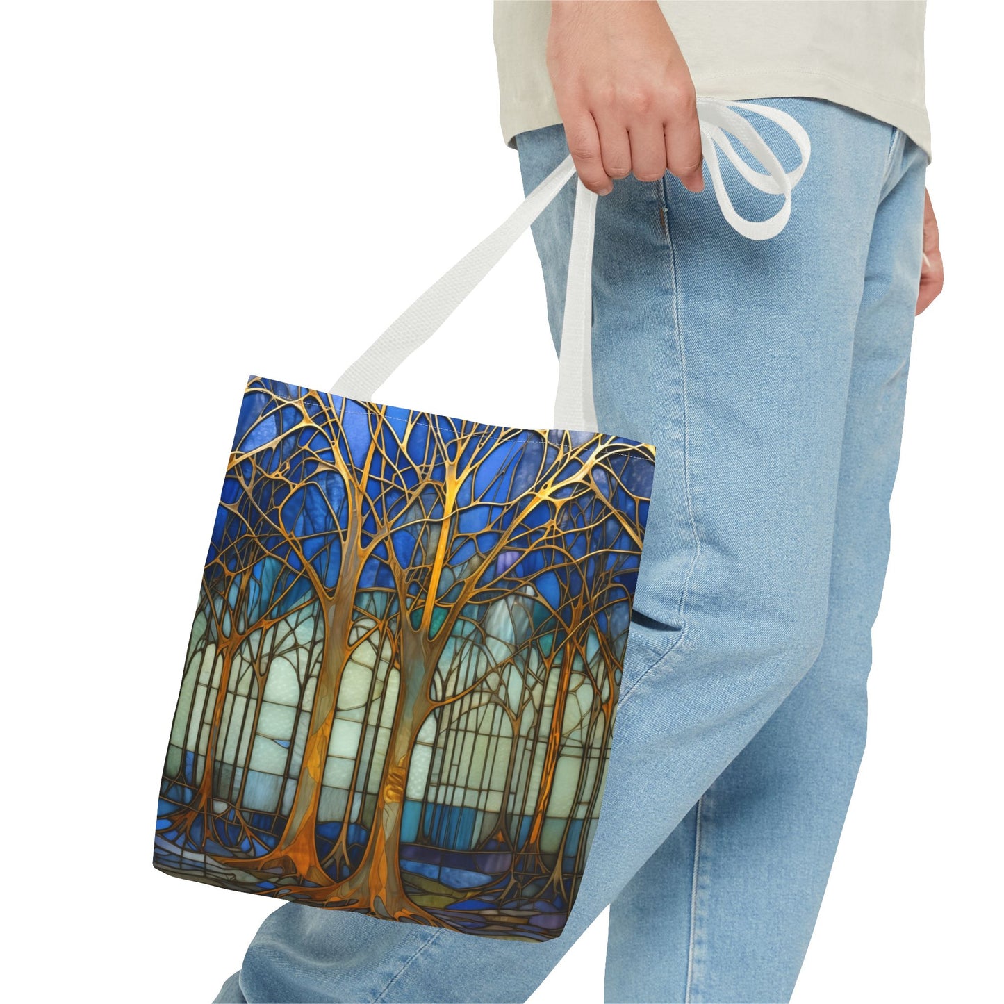 A person in jeans holds the Stained Glass Forest Tote Bag, showcasing an original artist design with trees on a vibrant blue background. Available in 3 sizes.