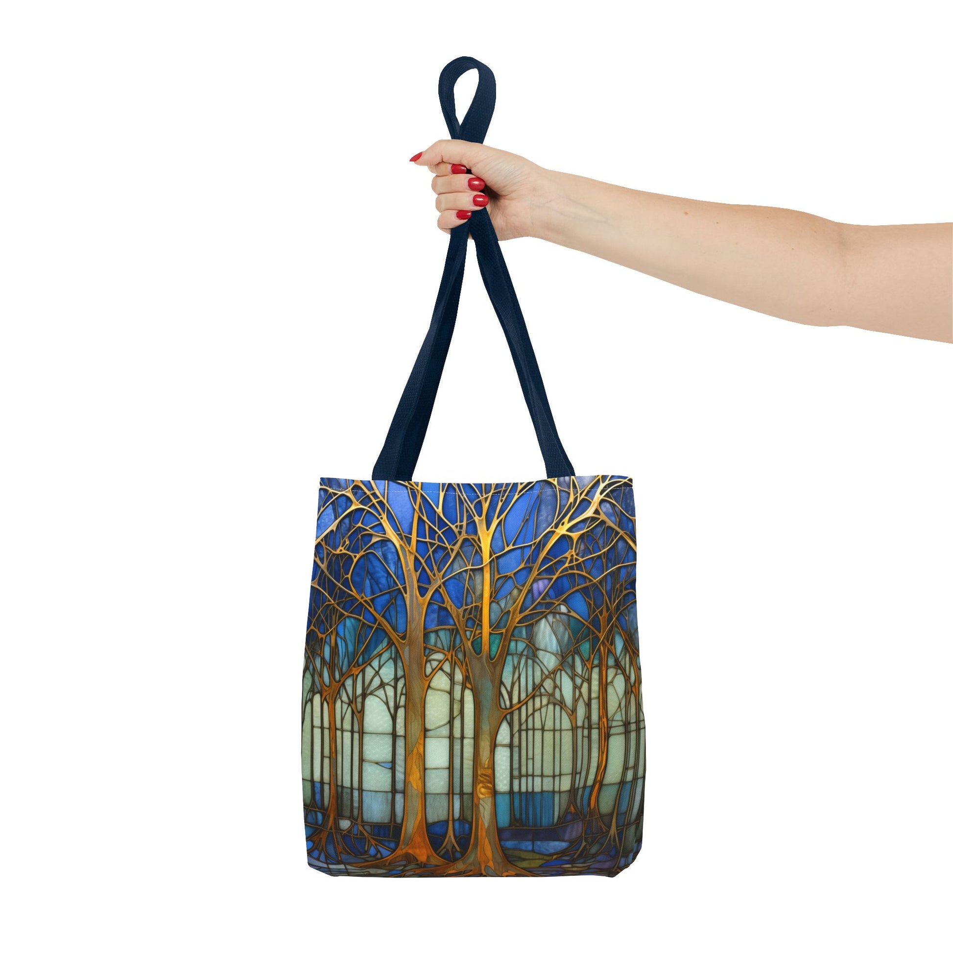 A person holding the Stained Glass Blue Forest Tote Bag with black straps features a vibrant forest design with gold trees against a blue and white background. Crafted from durable polyester, this stylish and sturdy tote bag is perfect for your daily errands and comes in three different sizes.