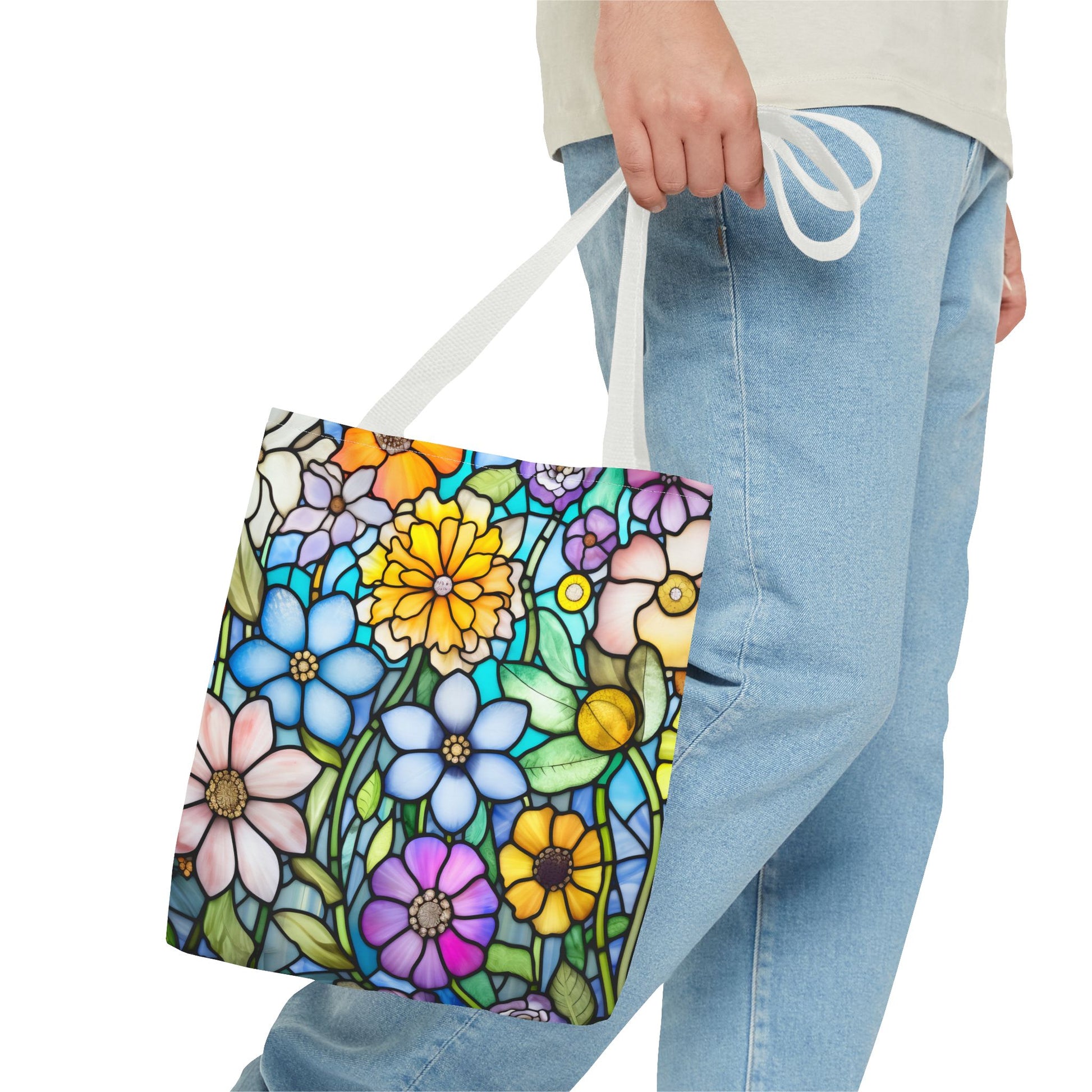 A person wearing denim jeans and a light shirt holds a Stained Glass Folk Art Flowers Tote Bag, adorned with a vibrant floral pattern and crafted from durable polyester materials.