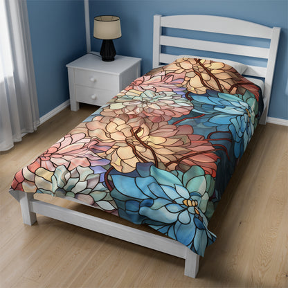 A bed adorned with the Stained Glass Flowers Soft Plush Blanket featuring large blue, pink, and yellow flowers rests against a blue wall. Beside it is a white nightstand with a lamp, and light wood flooring completes the cozy room setting.