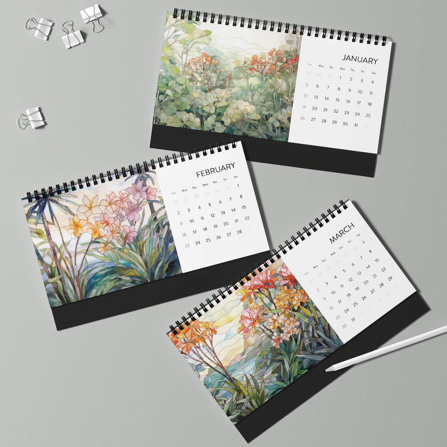 Three spiral-bound desk calendars for January, February, and March 2025 are laid out on a table, each featuring stained glass illustrations of tropical flowers. A pen and binder clips are placed nearby. These beautifully made in the USA desk calendars add a touch of elegance to any workspace.