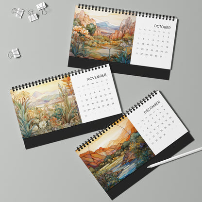 Three 2025 Desk Calendars - Desert Landscapes for October, November, and December are displayed on a light gray surface, each featuring scenic artwork on high-quality paper stock. A pen and two binder clips are also visible alongside the calendars.