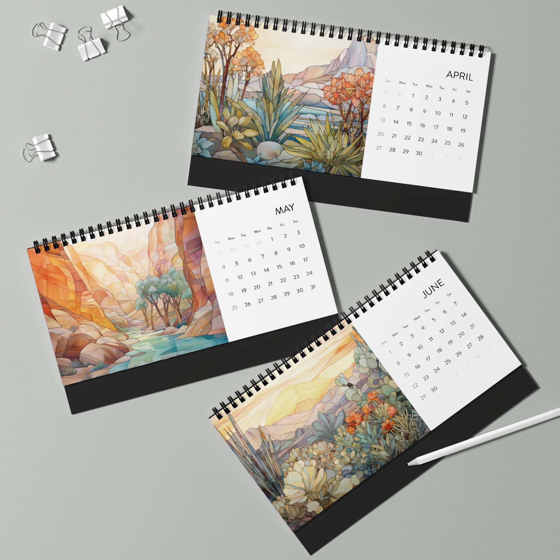 Three open 2025 Desk Calendars - Desert Landscapes, showcasing April, May, and June with watercolor-like nature scenes on high-quality paper stock. Nearby are three binder clips and a white pen on a light gray surface. The scenes evoke the beauty of stained glass illustrations, adding elegance to your workspace.