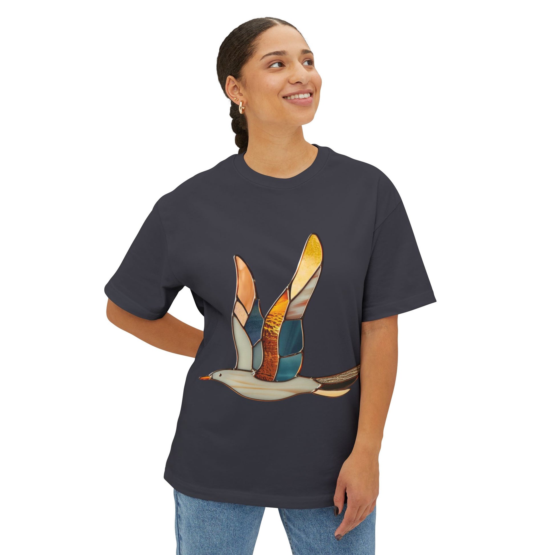 A person with a ponytail wears an oversized sustainable T-shirt from Bella+Canvas, featuring a colorful stained glass-style seagull design. They are looking up and smiling, with one hand in their pocket.