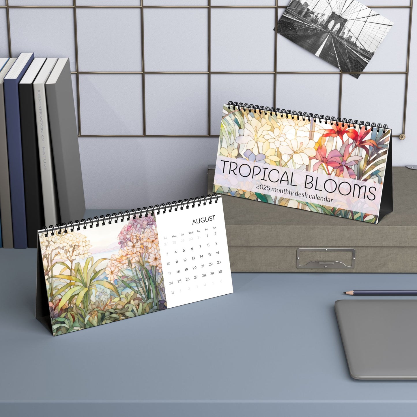 Two 2025 Desk Calendars - Tropical Flowers, proudly made in the USA, are displayed on a desk alongside books and a pen. One calendar is open to August, featuring stained glass illustrations of tropical flowers, while the other displays its front cover.