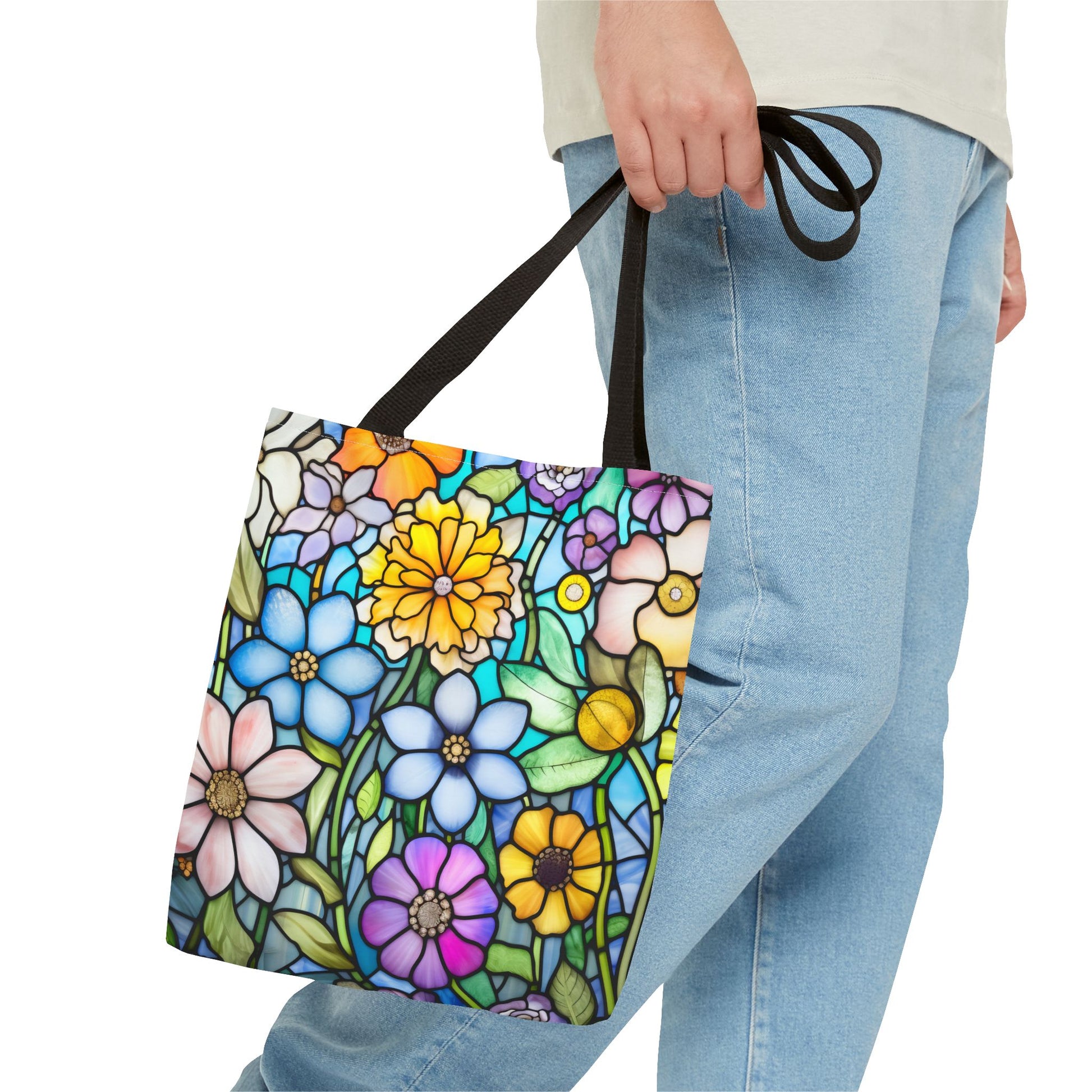 Someone holds a Stained Glass Folk Art Flowers Tote Bag featuring vibrant blue, yellow, pink, and purple floral designs. Made from durable polyester, it pairs seamlessly with their light jeans and shirt. Available in 3 sizes.