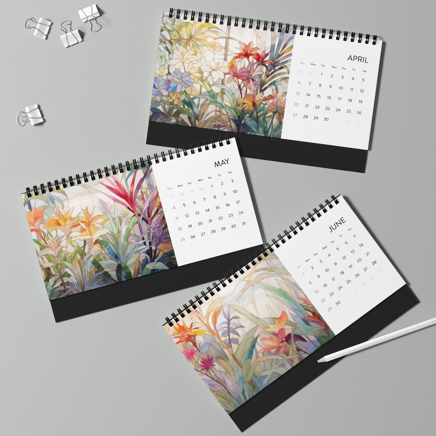 On a light grey surface, a white pen and binder clips are placed next to three spiral-bound 2025 Desk Calendars - Tropical Flowers for April, May, and June, each showcasing vibrant floral artwork.