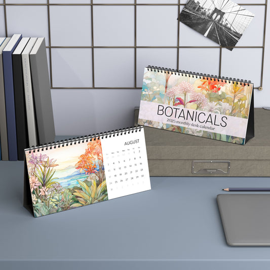 Two spiral-bound desk calendars titled "2025 Desk Calendar - Botanicals" are displayed on a desk next to books, a pencil, and a laptop. The calendar open to August showcases stunning botanical floral illustrations. Crafted with high-quality paper stock, these calendars are perfect for any workspace.