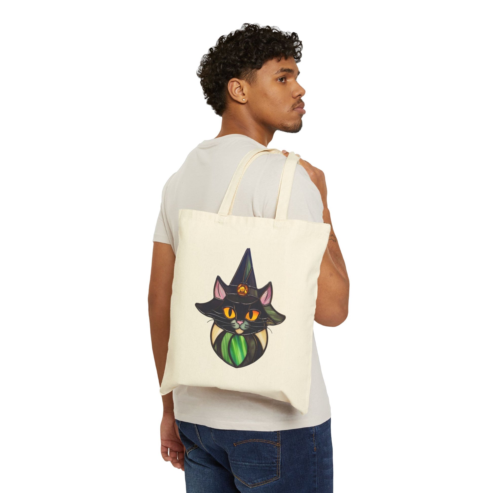 A person with curly hair is standing sideways, holding the "Stained Glass Cat Witch Cotton Canvas Tote Bag." They are wearing a light gray t-shirt and blue jeans.