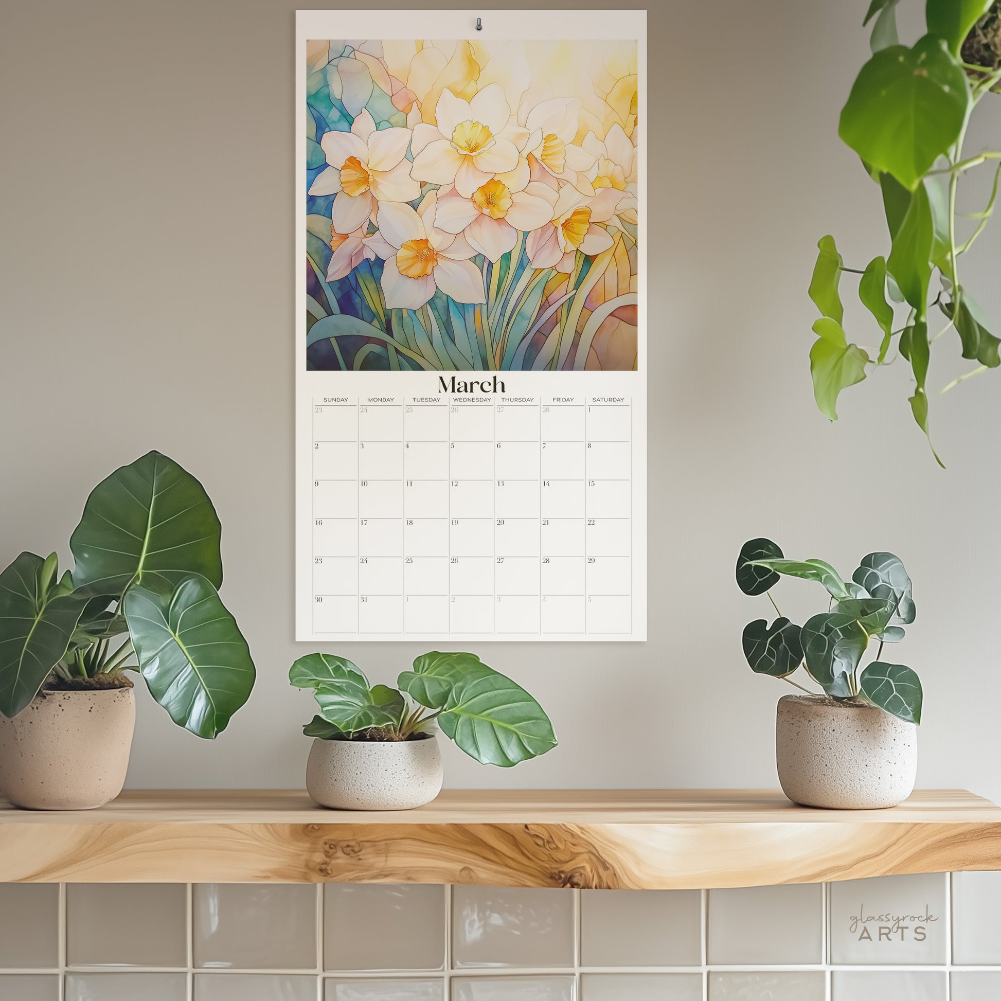 The birth flowers calendar, a 2025 monthly wall calendar with floral-themed original artwork, hanging on a wall surrounded by plants. The March page shows colorful daffodils.