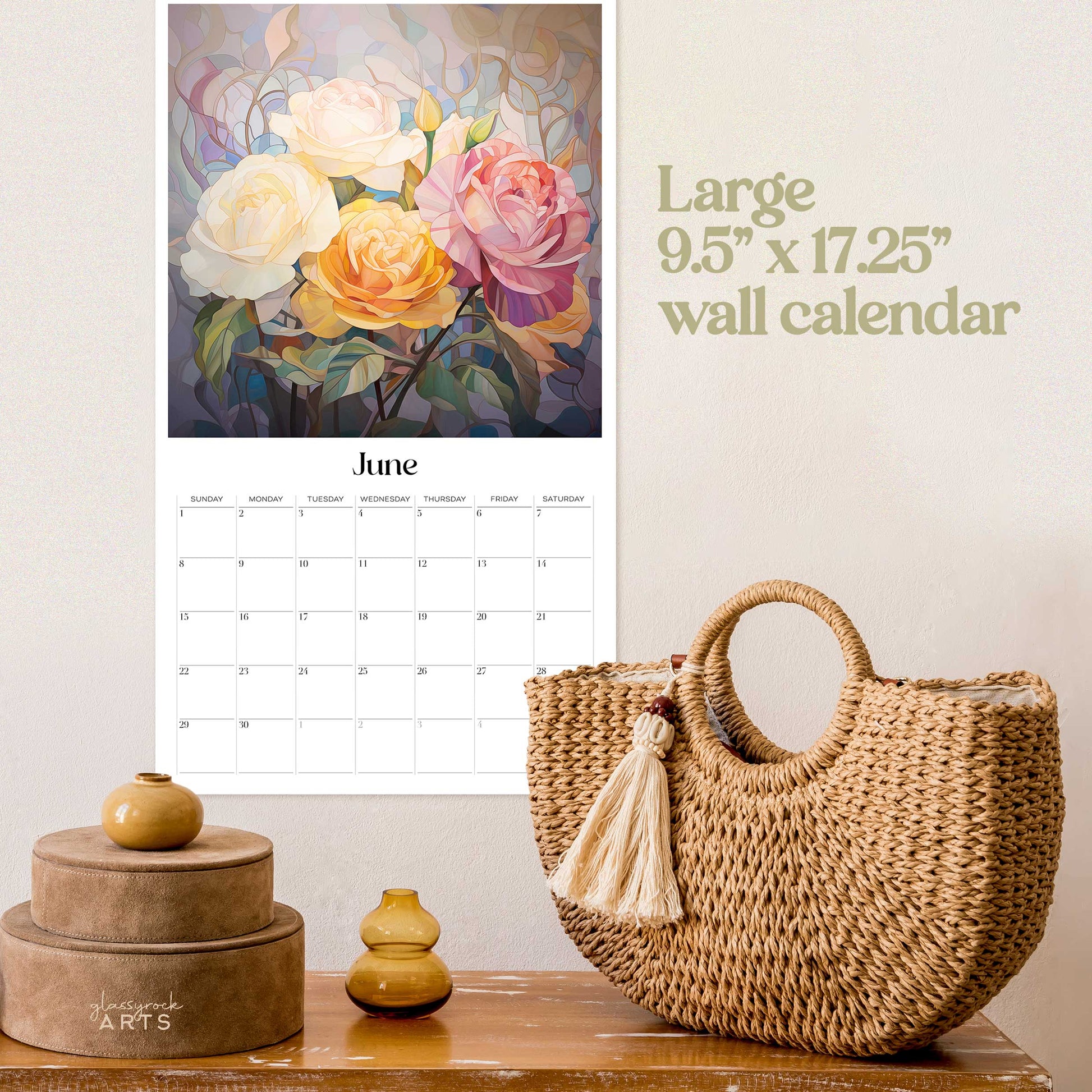 The birth flowers calendar, a 2025 monthly wall calendar with flower-themed original artwork, on a wall above a wood table with a wicker purse. The calendar is open to June, showing roses.