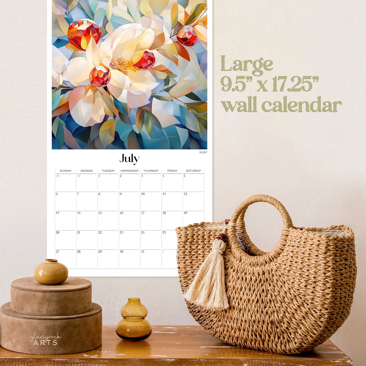 The birthstones calendar, a 2025 monthly wall calendar with gem-themed original artwork, on a wall above a wood table with a wicker purse. The calendar is open to July, showing rubies.