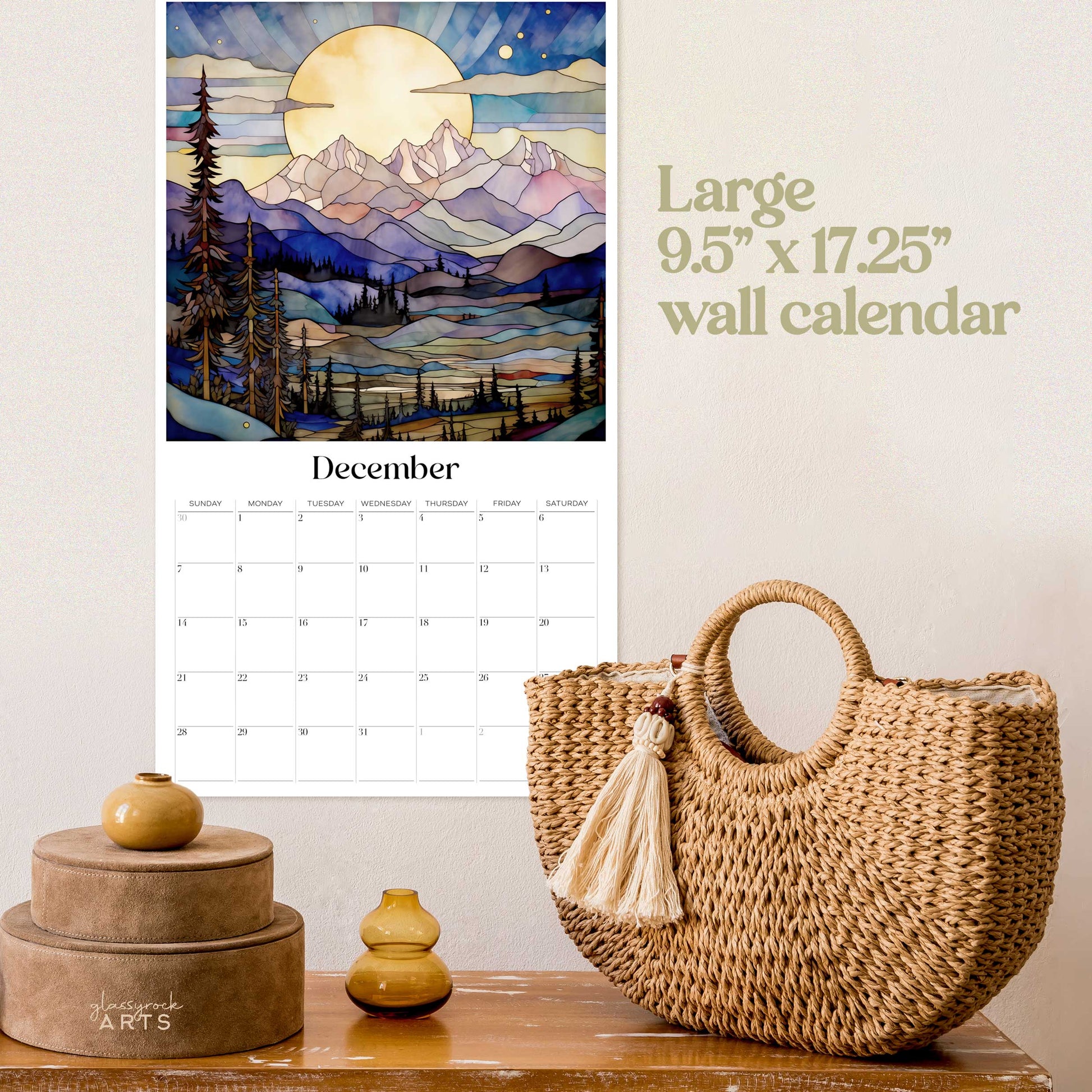 The 2025 stained glass mountains wall calendar on a beige wall with a wicker basket. The December page is showing. 