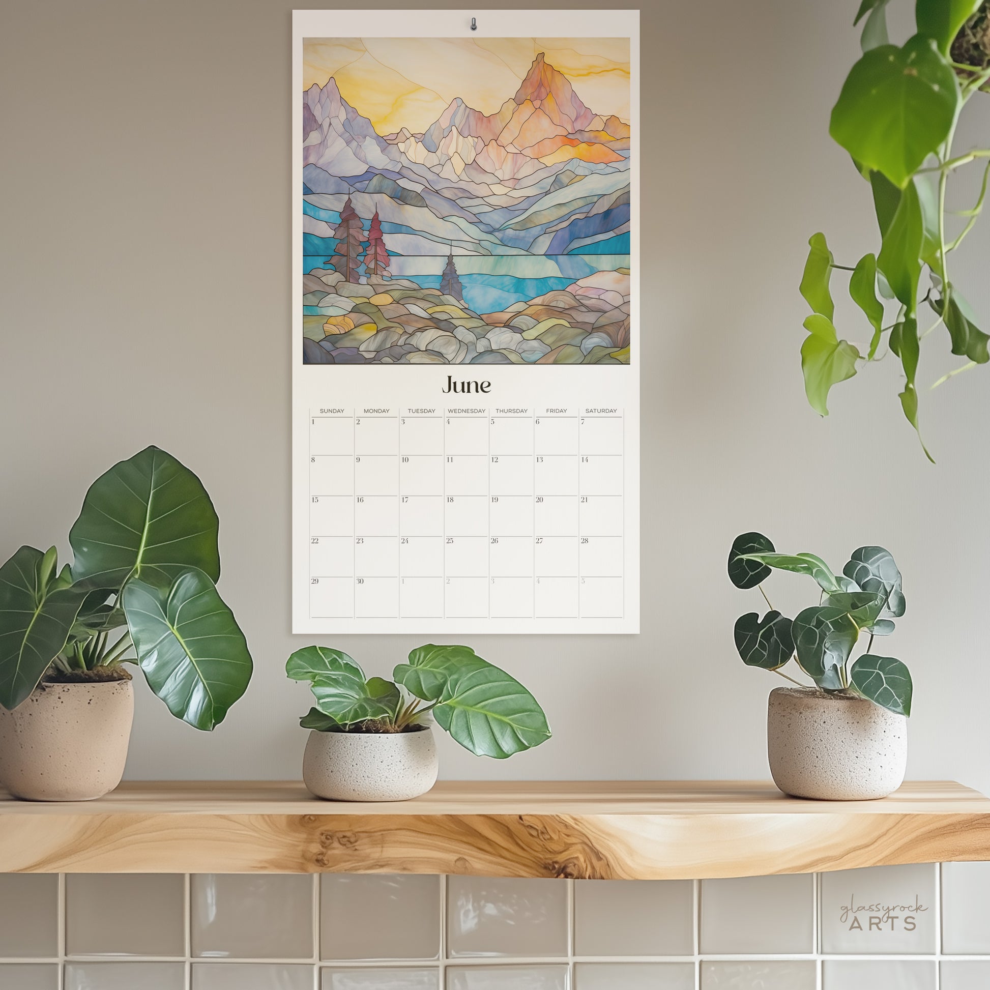 The 2025 stained glass mountains wall calendar on a grey wall with plants. The June page is showing. 