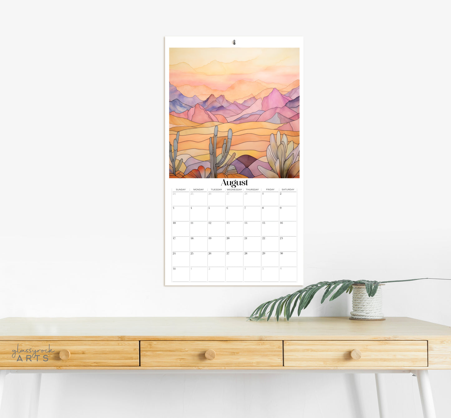 The 2025 stained glass mountains wall calendar on a grey wall with a desk. The August page is showing. 