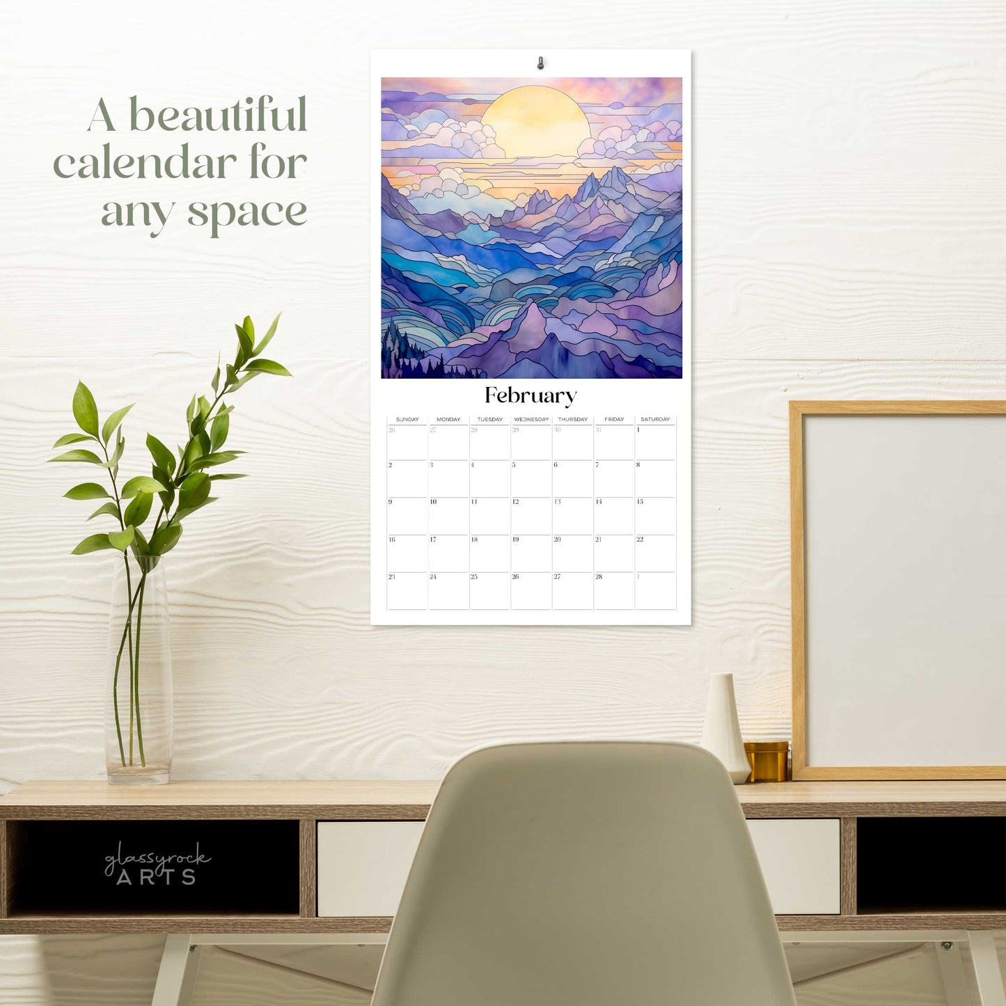 The 2025 stained glass mountains wall calendar on a grey wall with plants and a desk. The February page is showing. 