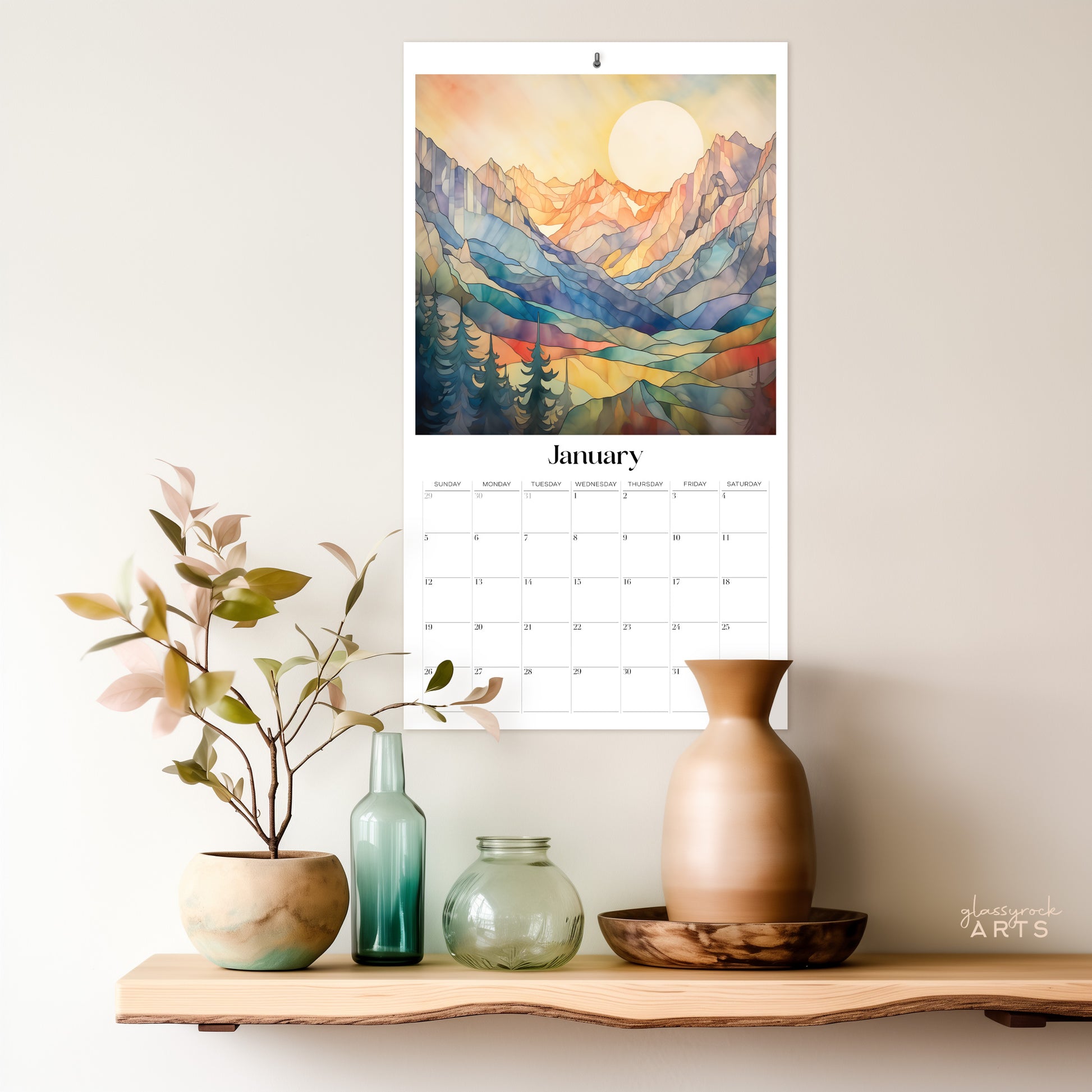 The 2025 stained glass mountains wall calendar on a grey wall with a natural edge wood shelf. The January page is showing. 