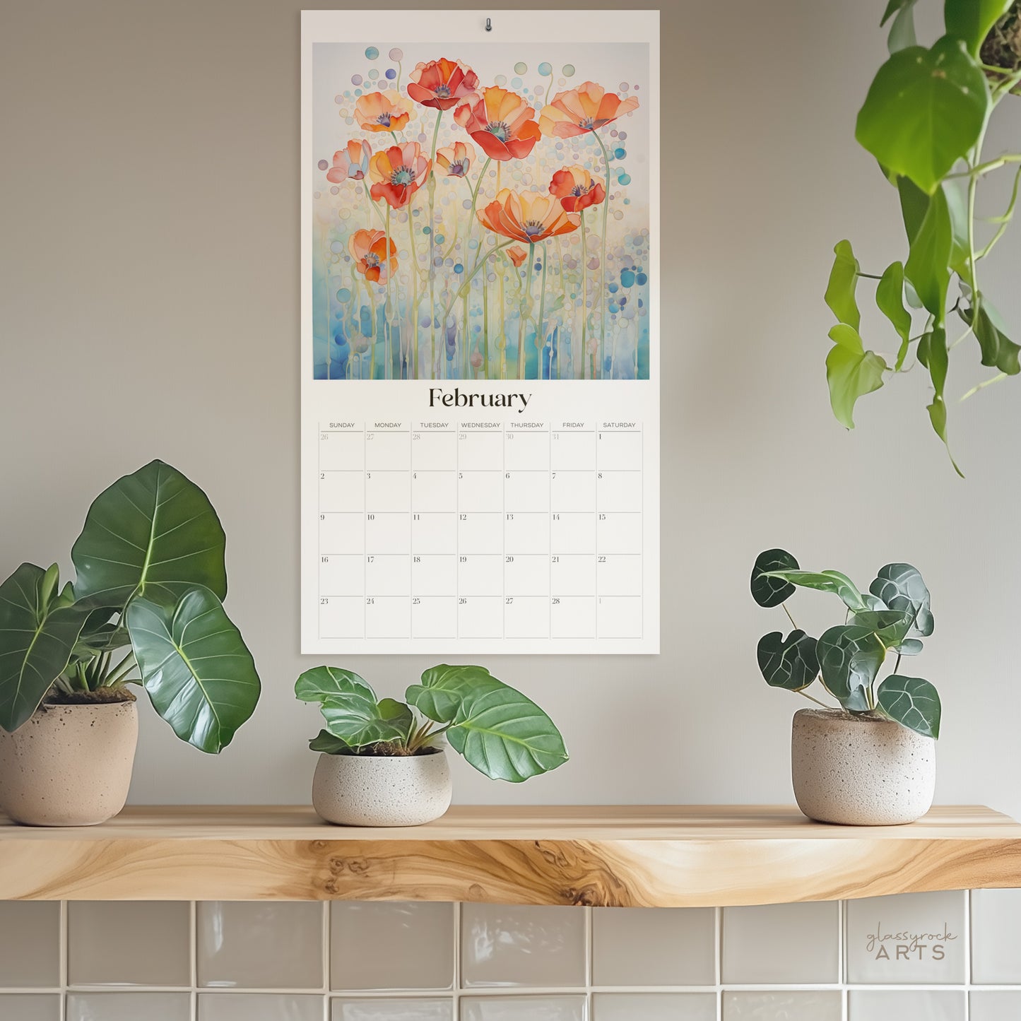 The 2025 Poppies Wall Calendar for February displays vibrant orange poppies, reminiscent of stained glass. It hangs above a wooden shelf with three potted plants against a softly muted wall featuring lush green foliage.