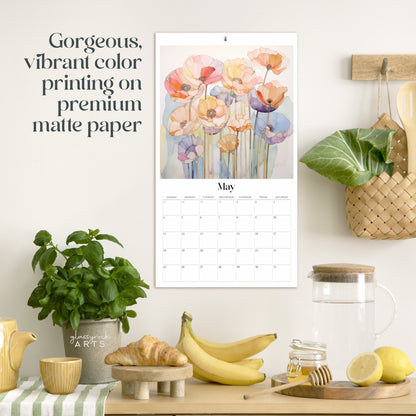 A kitchen scene displaying a 2025 Poppies Wall Calendar with vibrant May flowers on premium matte paper, evoking stained glass artistry. Below are a plant, striped towel, bananas, and glass jars.