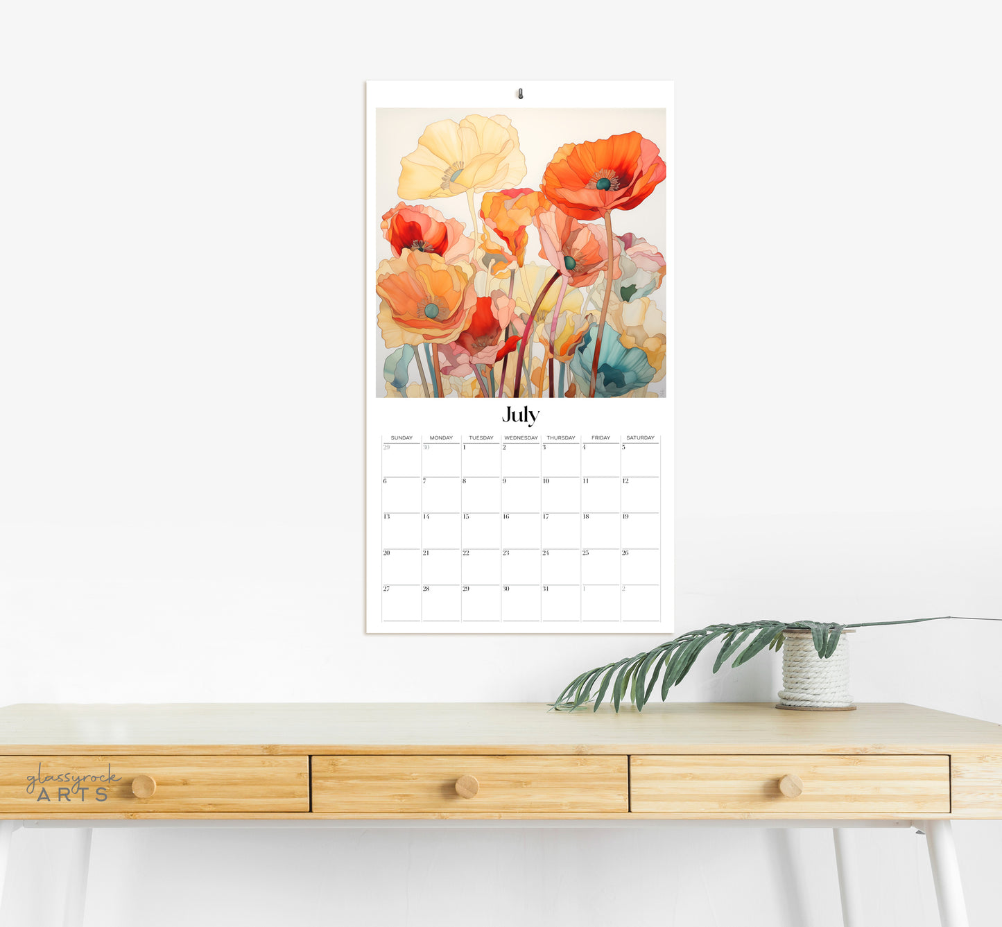 The 2025 Poppies Wall Calendar for July, adorned with vibrant flowers like stained poppies, is displayed on a white wall above a light wooden desk. A small potted plant rests on the desk, ensuring a clear and minimalist surface.