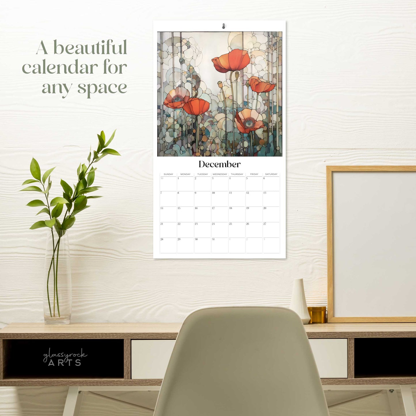 The 2025 Poppies Wall Calendar, featuring Decembers stained poppies artwork, hangs above a desk adorned with a plant, candle, and frames. Text on the wall reads, A beautiful calendar for any space.