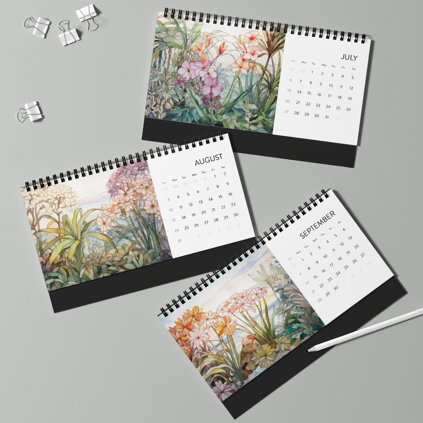 Three spiral-bound 2025 Desk Calendars - Tropical Flowers for July, August, and September feature botanical illustrations, displayed on a light gray surface with binder clips and a pen nearby. Made in the USA.