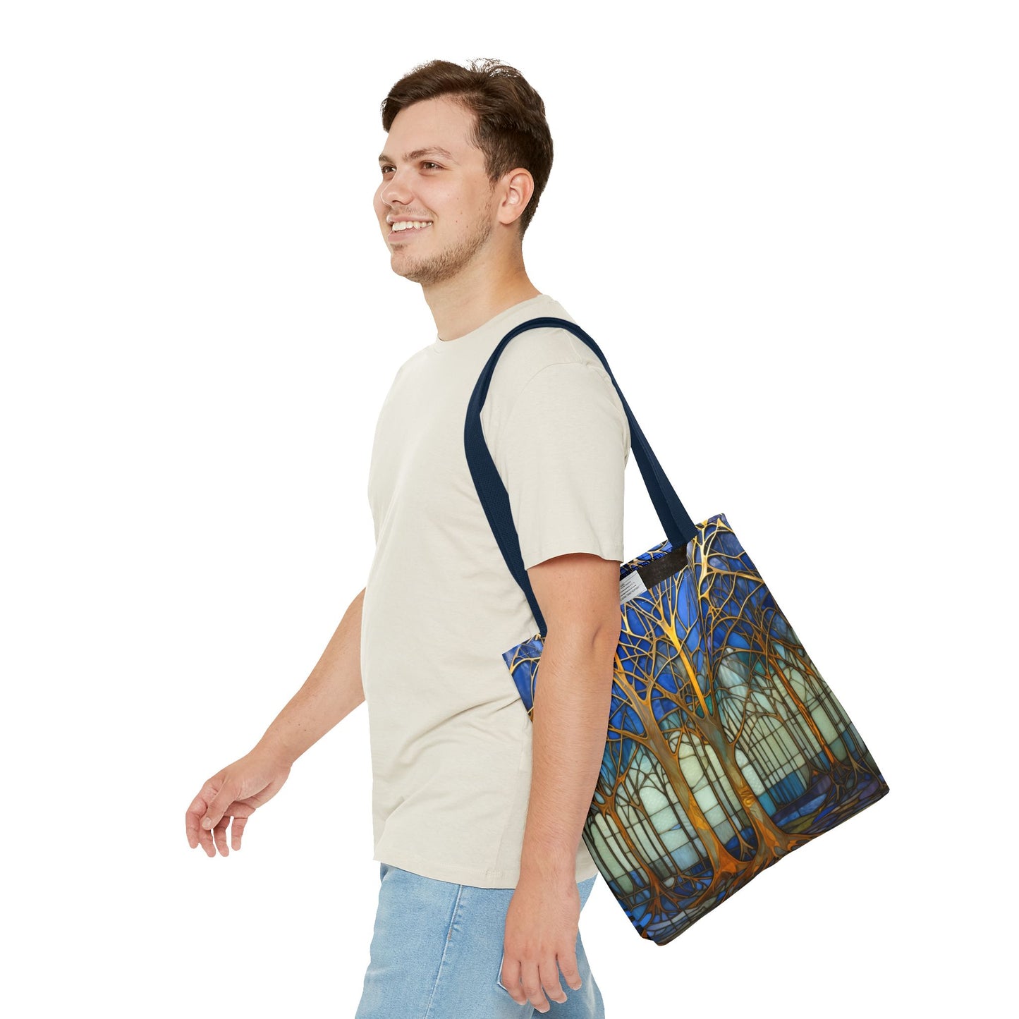 A person in a beige t-shirt and light blue jeans smiles while walking, carrying a Stained Glass Forest Tote Bag, Blue edition. The tote bag features an artistic tree and nature design set against a plain white background.