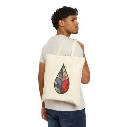 A person with short curly hair wears a light gray t-shirt and blue jeans, effortlessly slinging the cream Stained Glass Raindrop Cotton Canvas Tote Bag, showcasing an abstract leaf design, over their shoulder. The bag complements their side-turned pose against a plain backdrop.