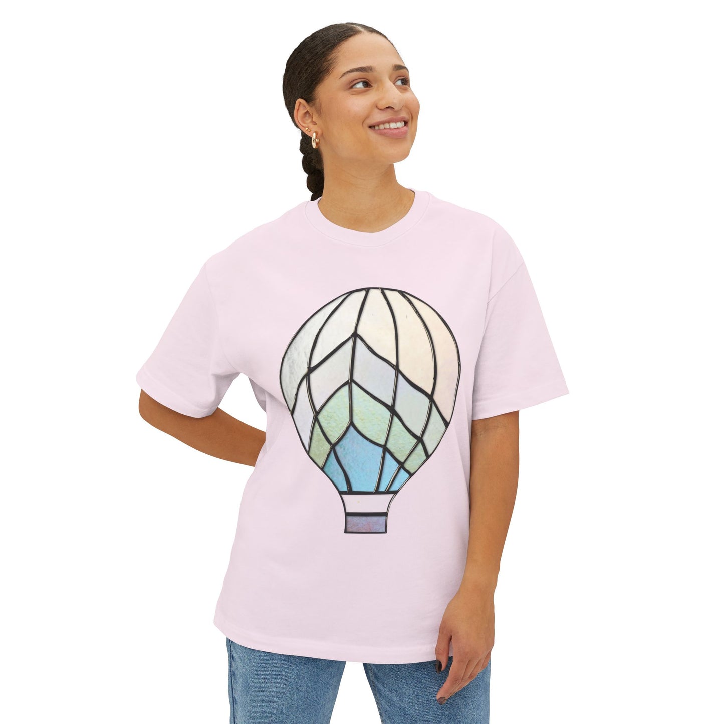 A person stands smiling, wearing the Stained Glass Hot Air Balloon Oversized Tee Shirt from Bella+Canvas, which features a light pink color and a large, stylized hot air balloon graphic on the front.