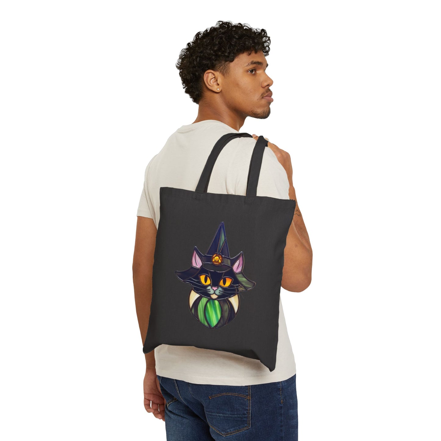 A person with curly hair wearing a white t-shirt and jeans, holding the Stained Glass Cat Witch Cotton Canvas Tote Bag over their shoulder.