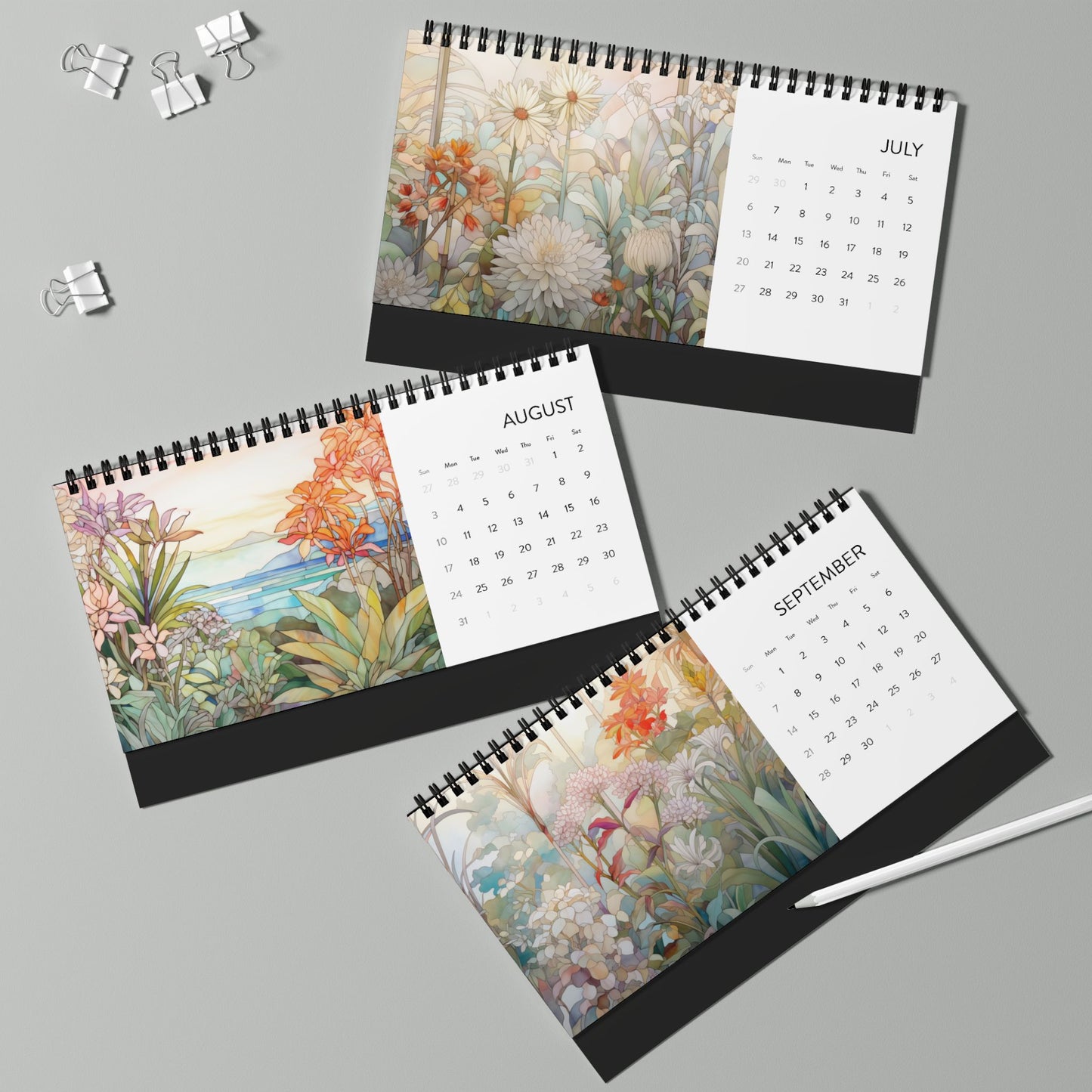 The 2025 Desk Calendar - Botanicals, crafted on high-quality paper stock, showcases the months of July, August, and September with botanical floral illustrations. Three metal binder clips and a white pen rest nearby on the light green surface.