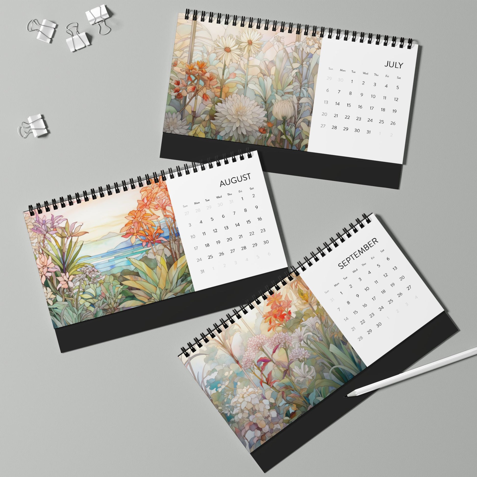 The 2025 Desk Calendar - Botanicals, crafted on high-quality paper stock, showcases the months of July, August, and September with botanical floral illustrations. Three metal binder clips and a white pen rest nearby on the light green surface.