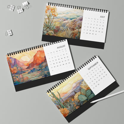 Three copies of the "2025 Desk Calendar - Desert Landscapes," each spiral-bound and printed on high-quality paper stock, are open to July, August, and September. Each calendar displays artistic stained glass landscape illustrations. A pen and two binder clips rest nearby on a grey surface.