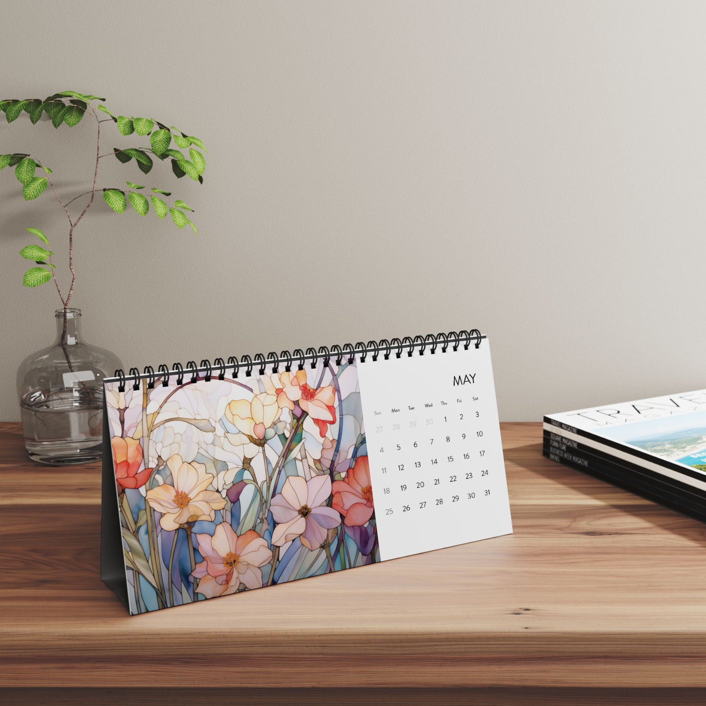 The 2025 Desk Calendar - Botanicals, showing the month of May with its botanical floral illustrations, stands on a wooden surface alongside a small plant and a stack of magazines, all printed on high-quality paper stock.