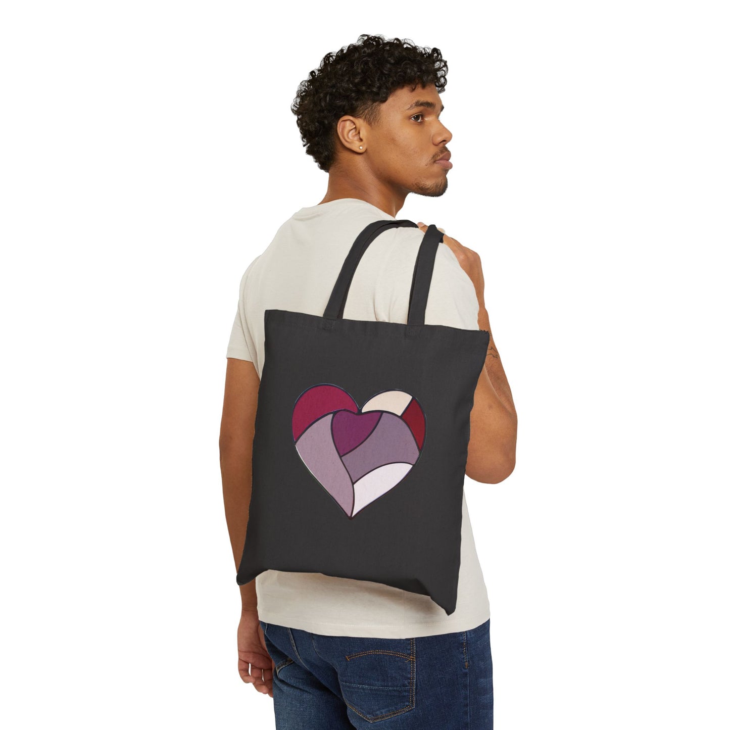 A person with short curly hair in a white T-shirt and jeans is seen from behind, holding the Stained Glass Heart Cotton Canvas Tote Bag. Designed by Noelle Barnes, this black tote features a colorful heart in purple and pink shades, resembling stained glass. The background is pristine white.