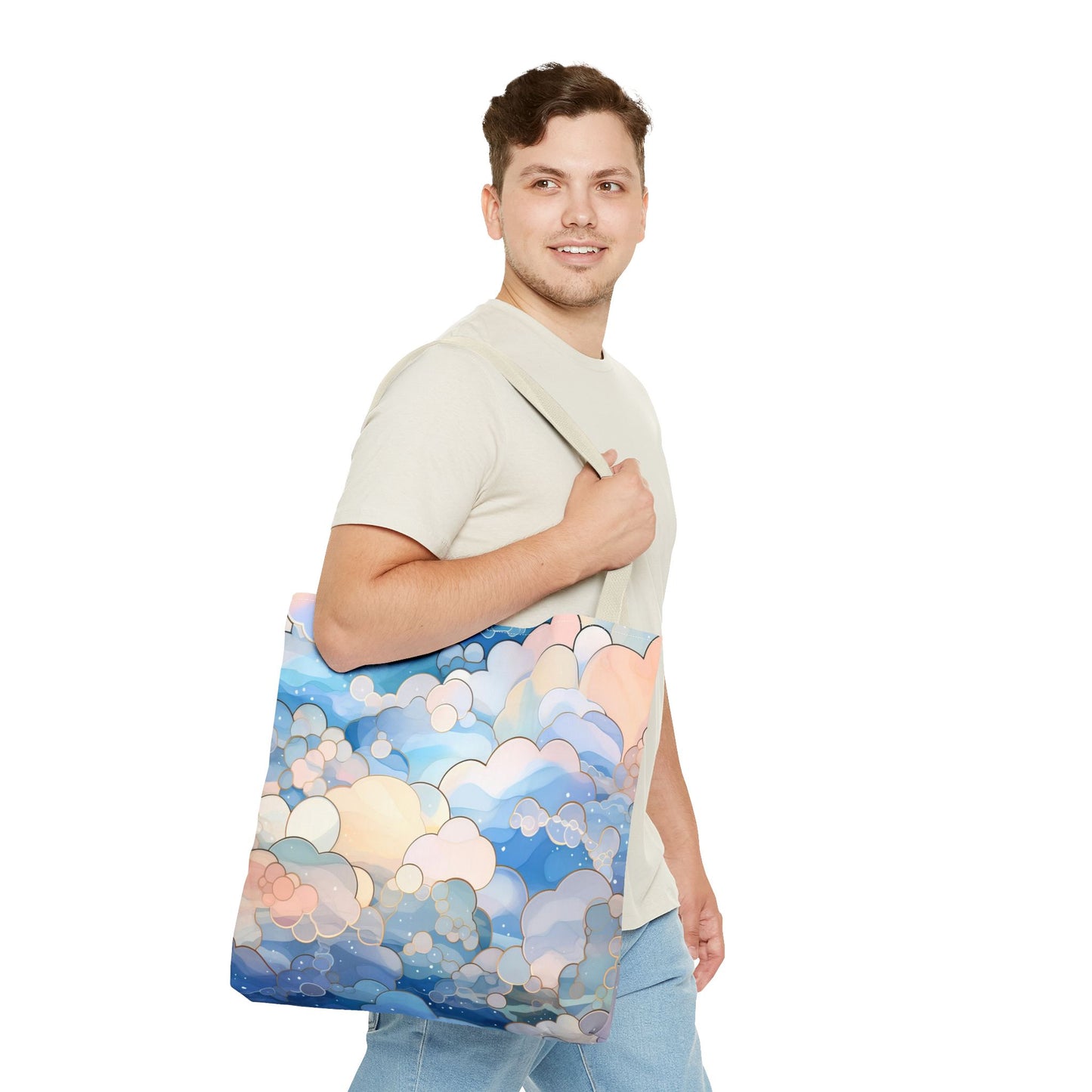 A person with short brown hair, wearing a light beige shirt and blue jeans, smiles while carrying a Stained Glass Clouds Tote Bag in blue. The durable polyester bag has an abstract cloud pattern with pink and peach tones. The background is plain white.