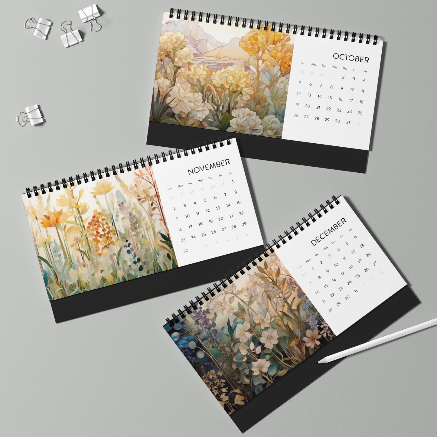 Three 2025 Desk Calendar - Botanicals, showcasing floral illustrations for October, November, and December on high-quality paper stock, are arranged on a light gray surface with binder clips and a white pen nearby.