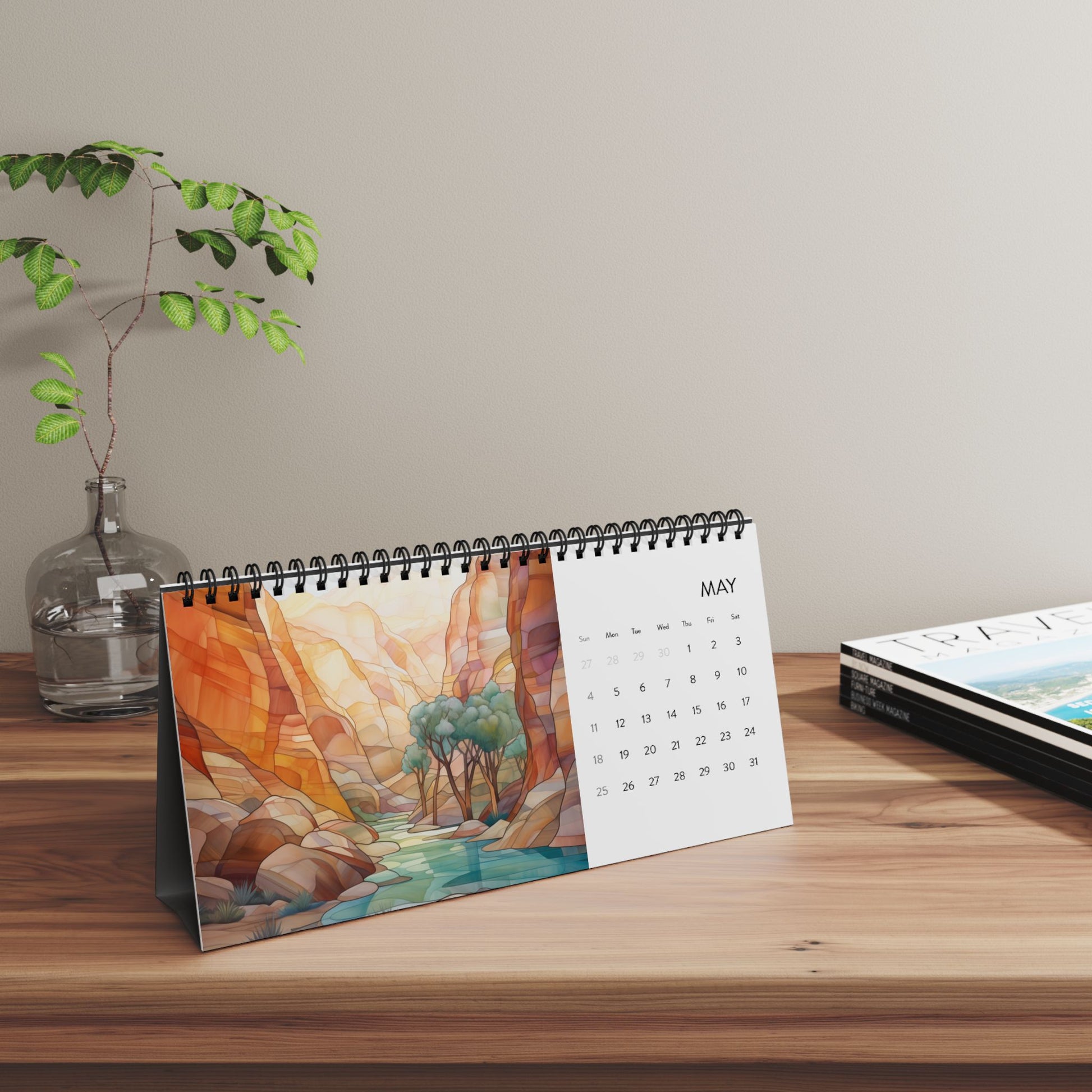 The 2025 Desk Calendar - Desert Landscapes, printed on high-quality paper stock and displaying the month of May with a vibrant canyon scene, is placed on a wooden surface next to a small potted plant and a stack of magazines.