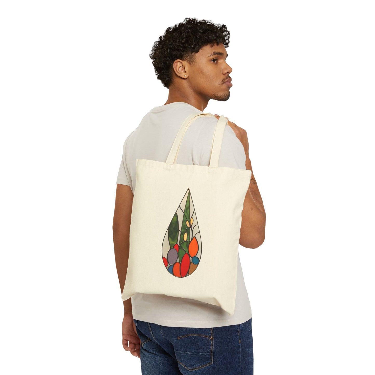 A person wearing a white T-shirt and jeans holds the Stained Glass Cactus Teardrop Cotton Canvas Tote Bag over their shoulder, showcasing its vibrant stained glass design of a teardrop with geometric shapes on a plain beige background, reminiscent of Noelle Barnes style.