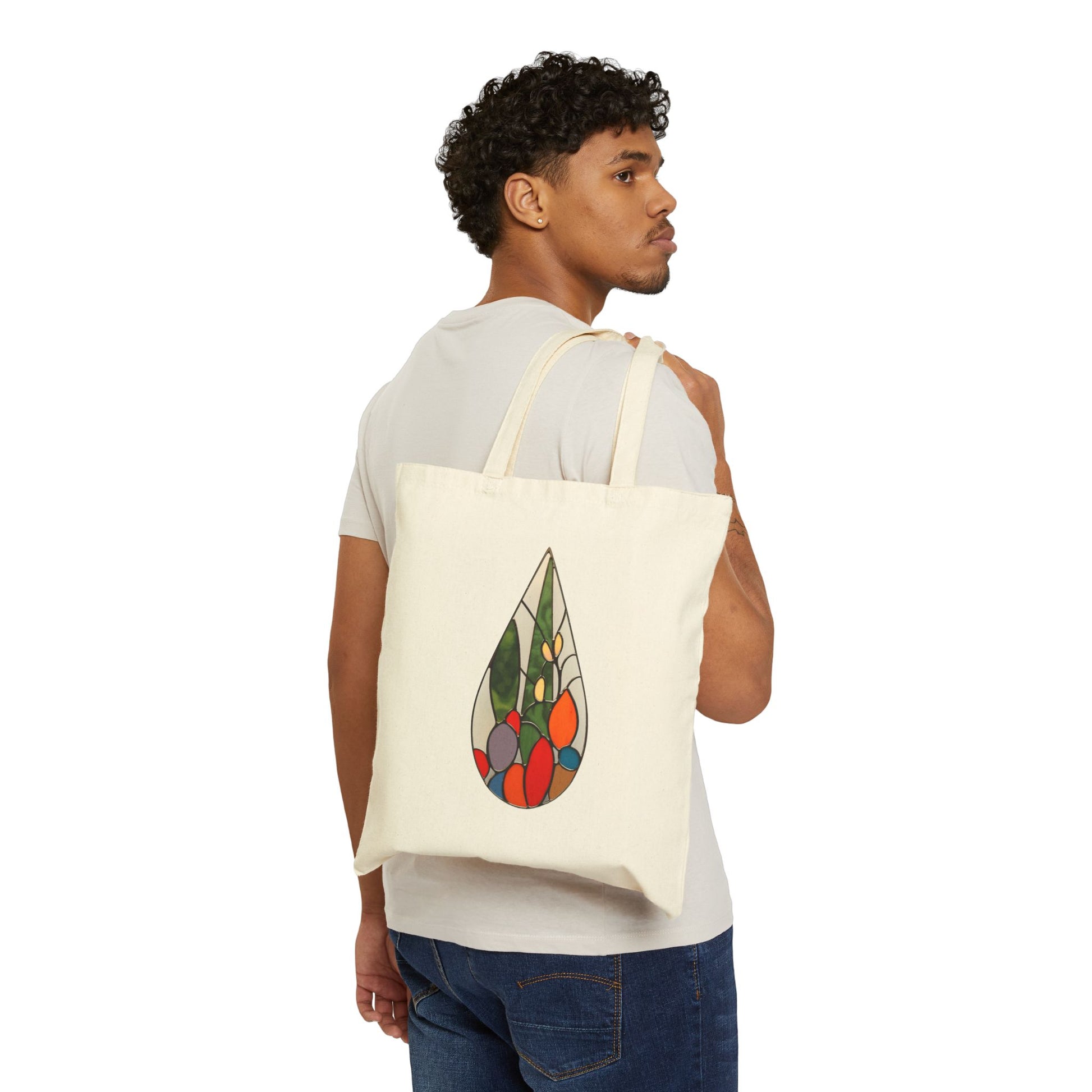 A person wearing a white T-shirt and jeans holds the Stained Glass Cactus Teardrop Cotton Canvas Tote Bag over their shoulder, showcasing its vibrant stained glass design of a teardrop with geometric shapes on a plain beige background, reminiscent of Noelle Barnes style.