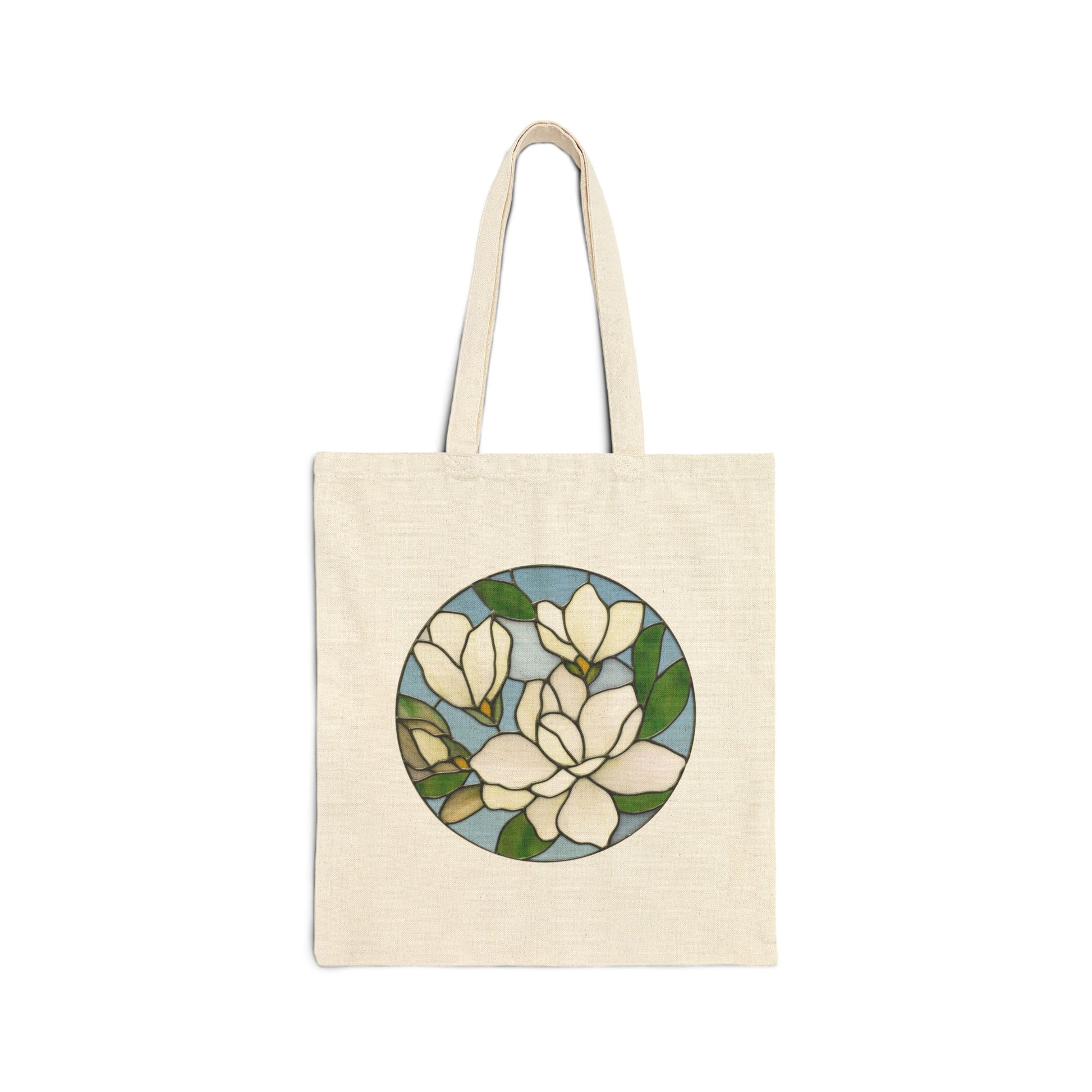 The Stained Glass Magnolia Cotton Canvas Tote Bag is a beige 100% cotton canvas tote bag with a circular floral stained-glass design featuring white magnolia flowers and green leaves on the front.